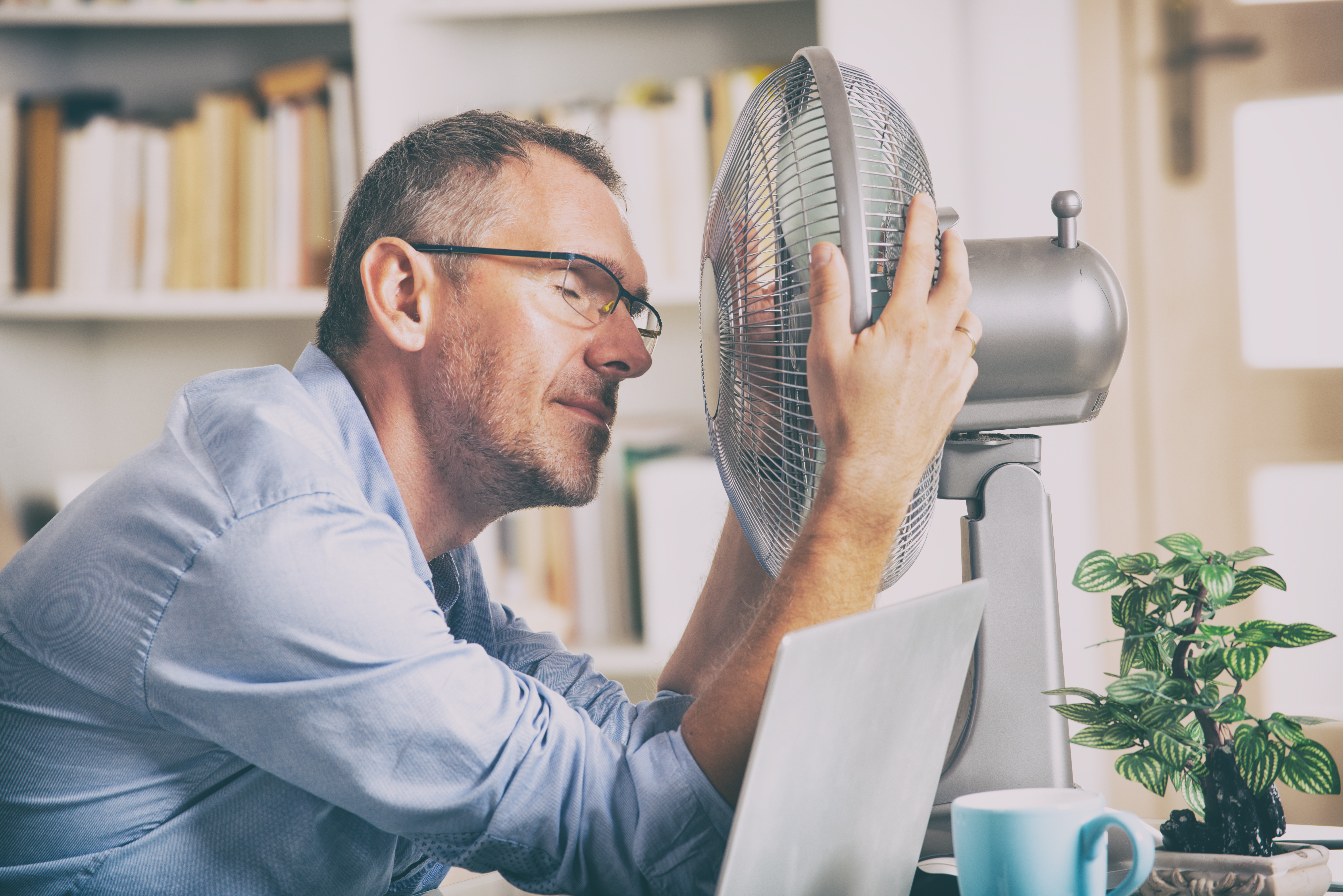 Mann sitzt im Büro vor einem Ventilator. Er hat die Augen zu und wirkt erschöpft.